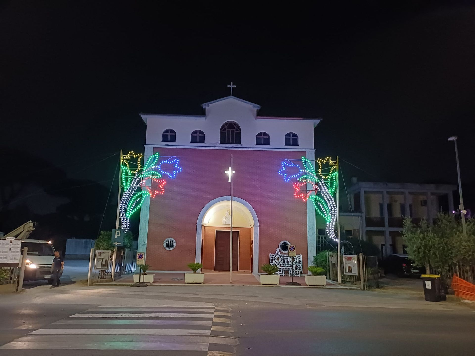 Una chiesa rosa di notte, decorata con luci a forma di fiore, caratterizzata da un portale ad arco e una croce in cima.