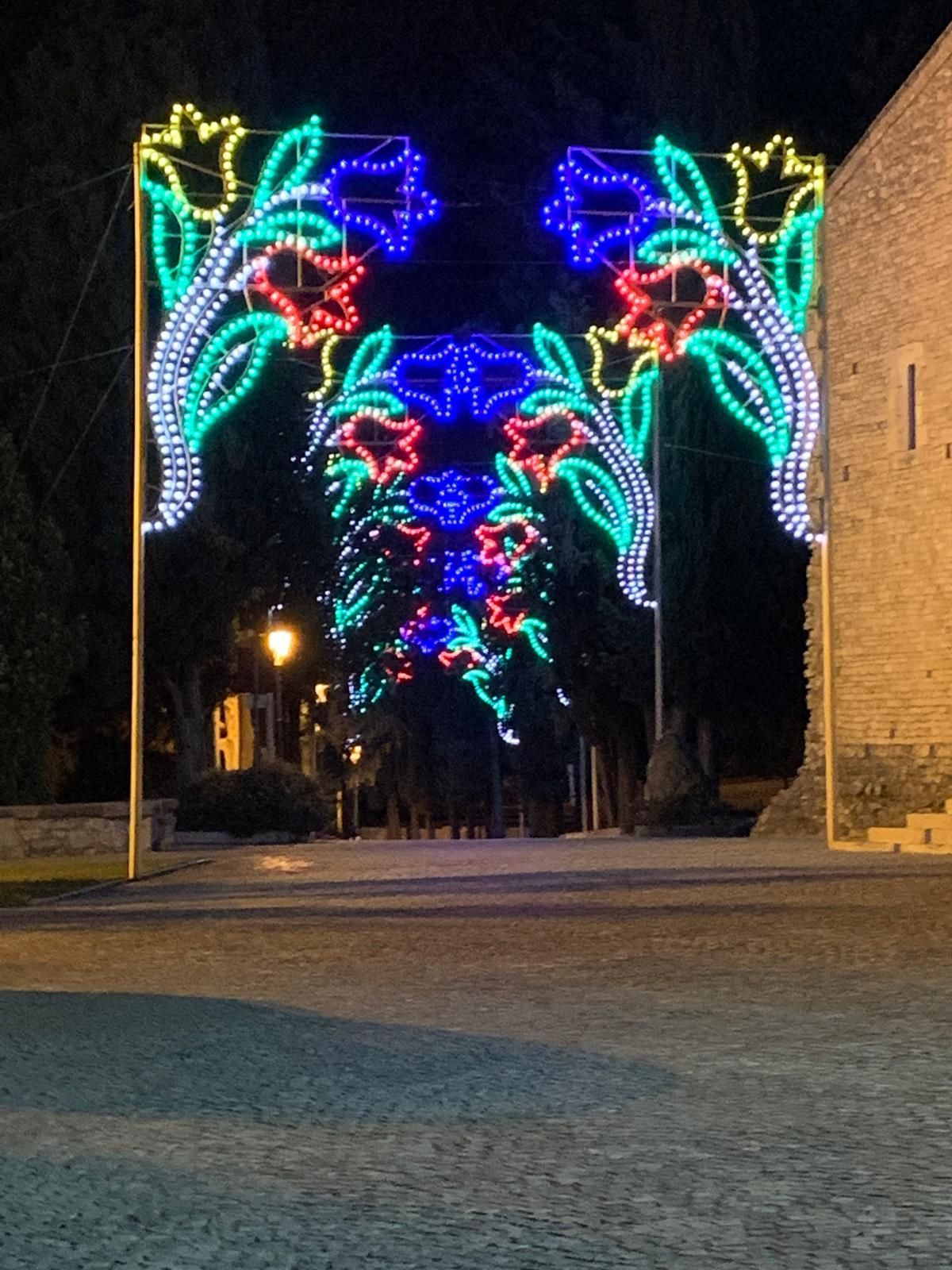 Un tunnel di luci LED colorate a forma di fiori illumina di notte un sentiero lastricato.