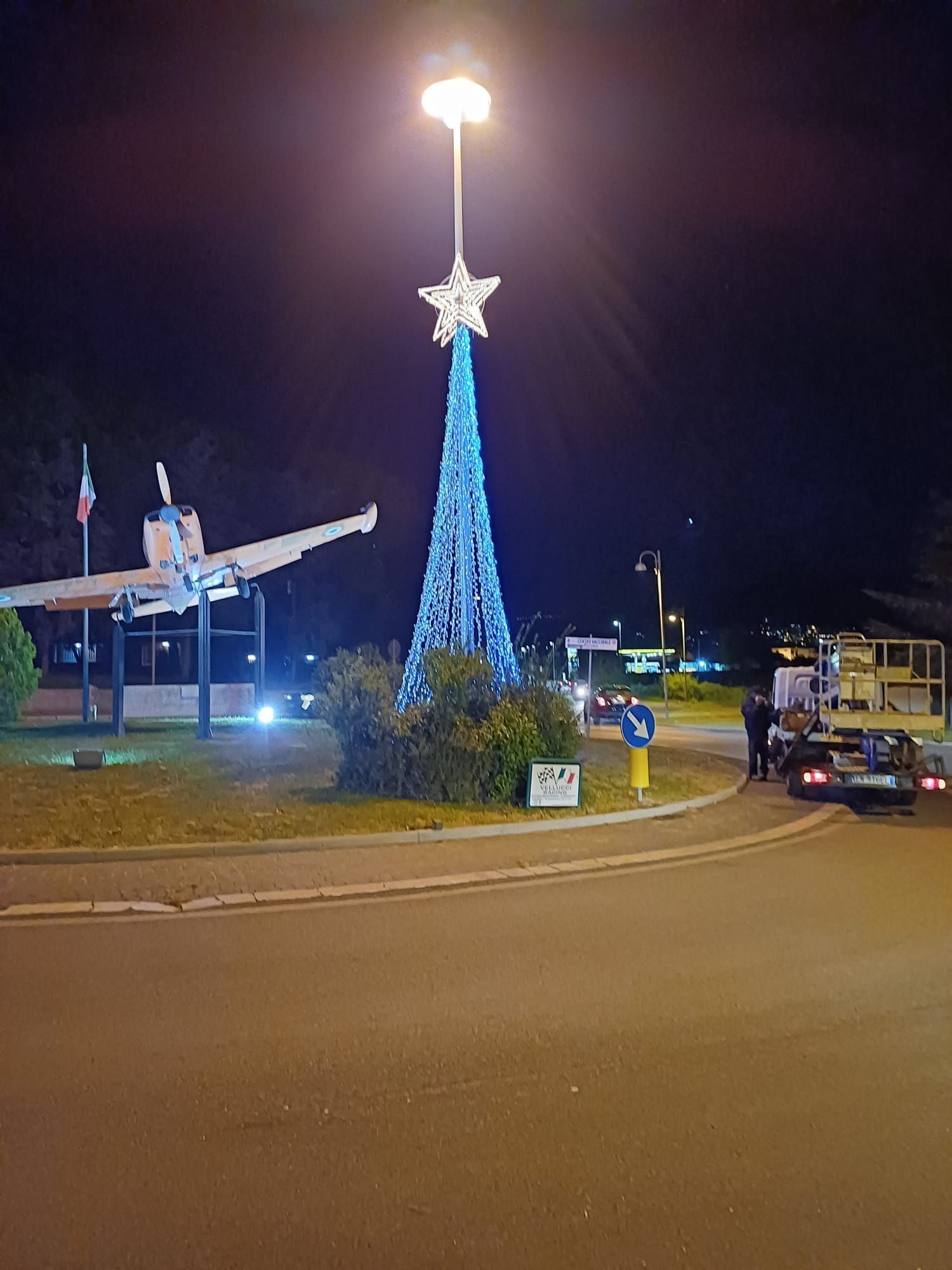 Una rotonda di notte con una scultura ad albero di Natale illuminata di blu, un aereo in mostra e veicoli nelle vicinanze.
