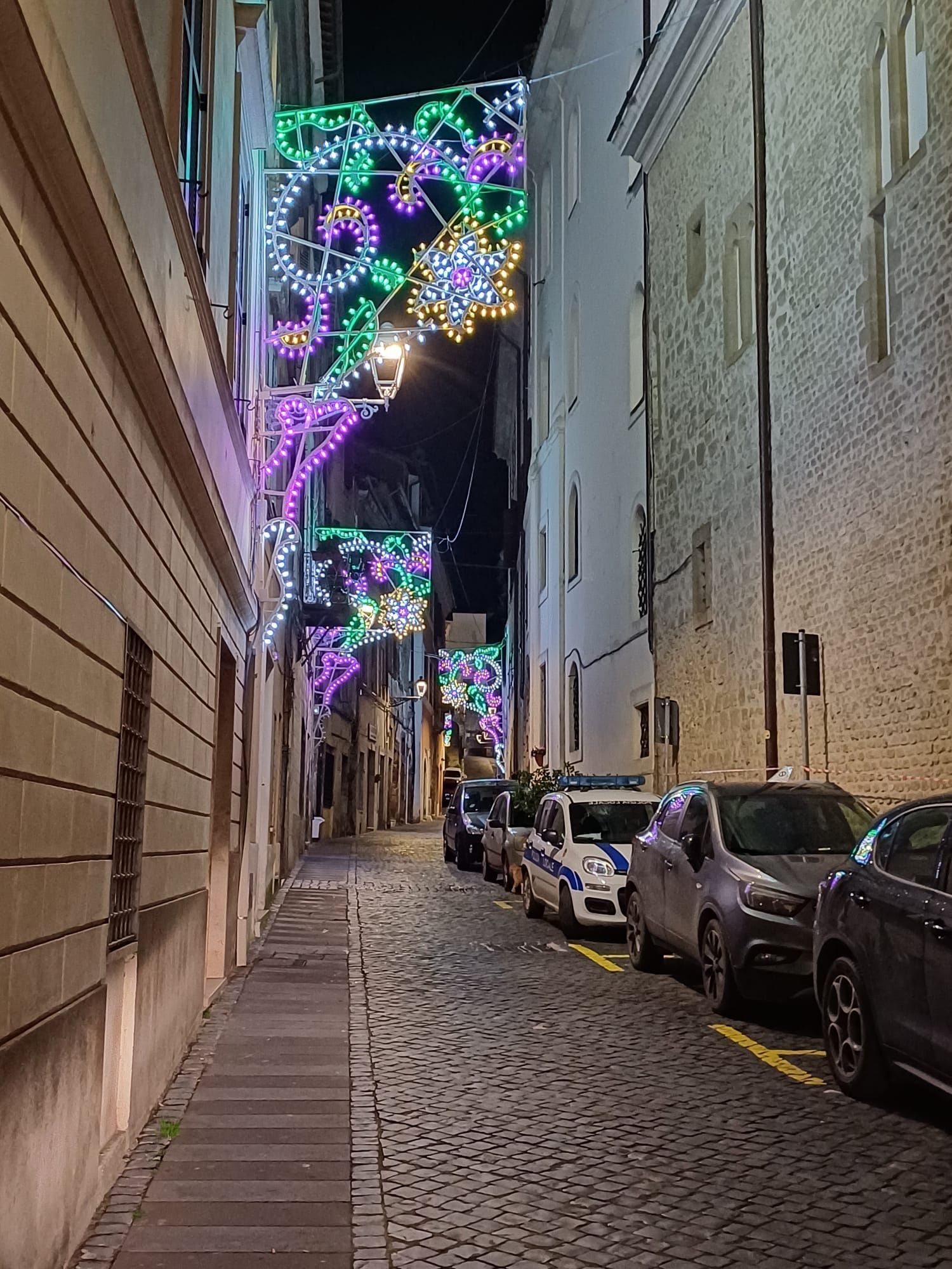 Una stretta strada europea lastricata di ciottoli, di notte, decorata con colorate e scintillanti luci natalizie sopra le auto parcheggiate.