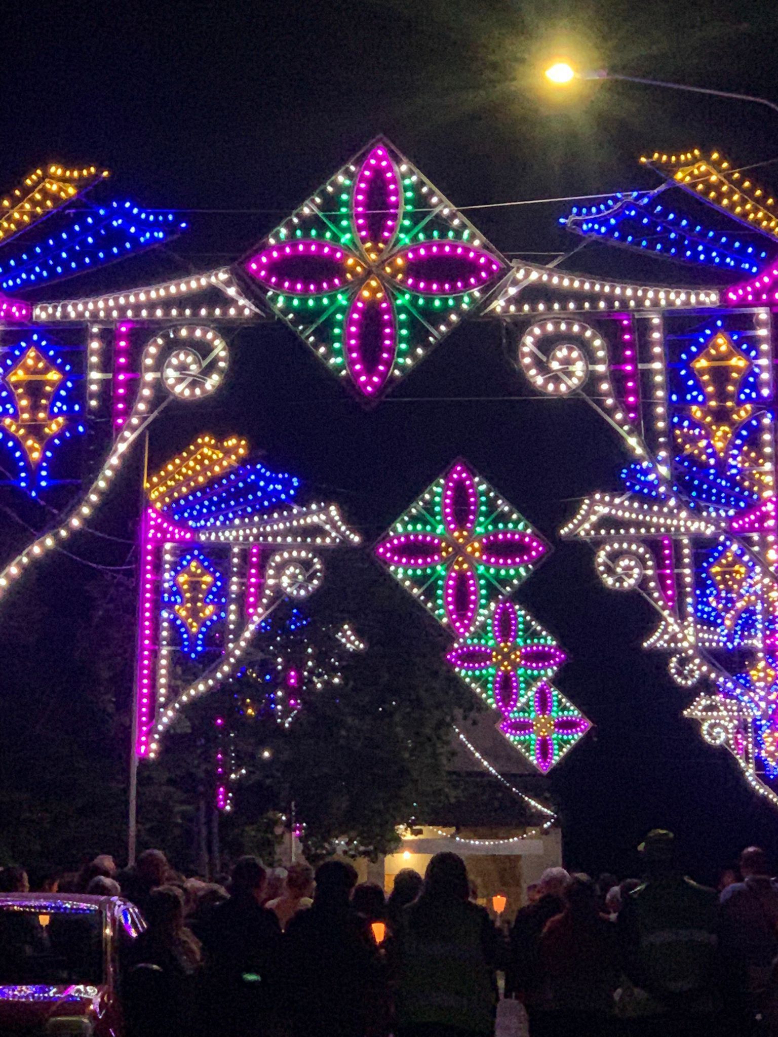 Un festoso arco adornato con colorate installazioni luminose geometriche illumina una folla di persone durante la notte.