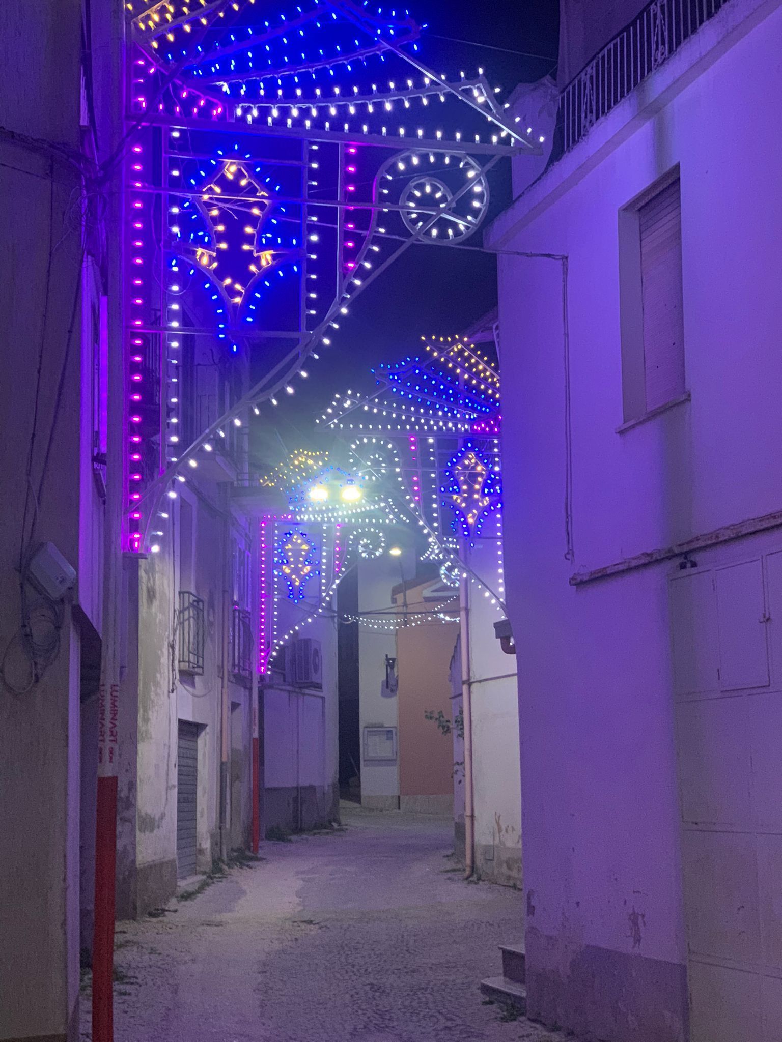 Una strada stretta illuminata di notte da vivaci luci natalizie decorative blu e viola, appese tra gli edifici.