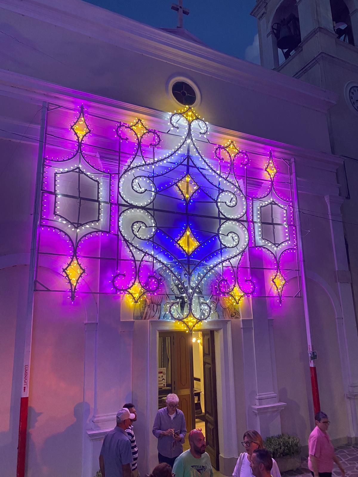 Al crepuscolo, alcune persone si trovano davanti alla facciata di una chiesa decorata con elaborate e luminose installazioni luminose viola e dorate.