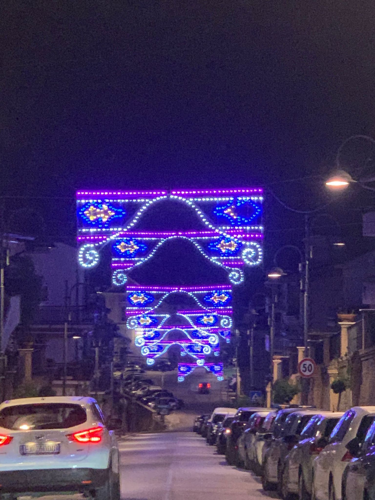 Una strada fiancheggiata da auto parcheggiate di notte, illuminata da una serie di archi luminosi decorativi viola e blu.