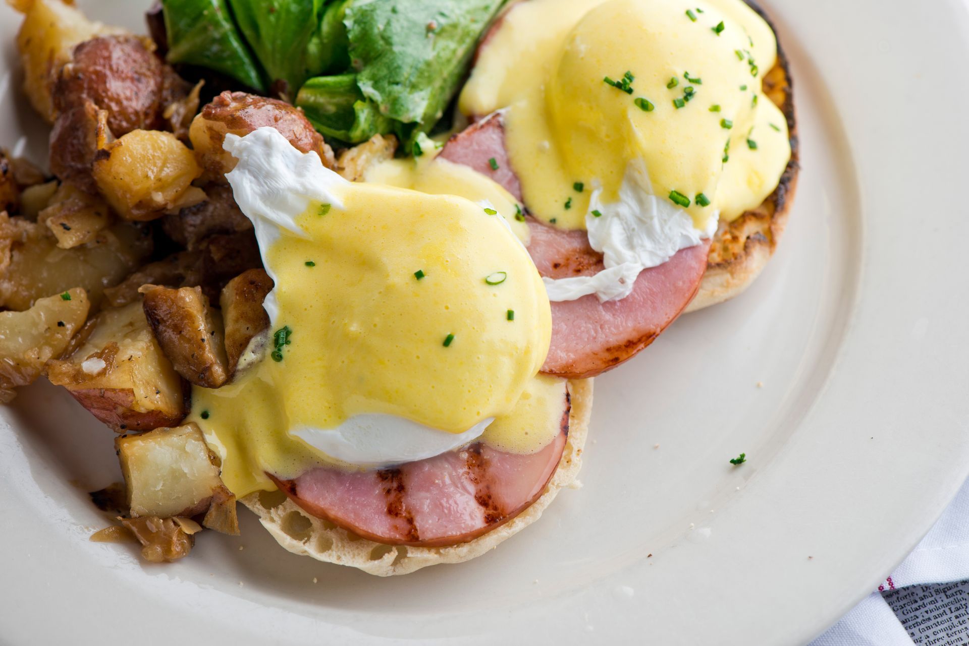Eggs Benedict with hollandaise, ham, roasted potatoes, and greens on a white plate