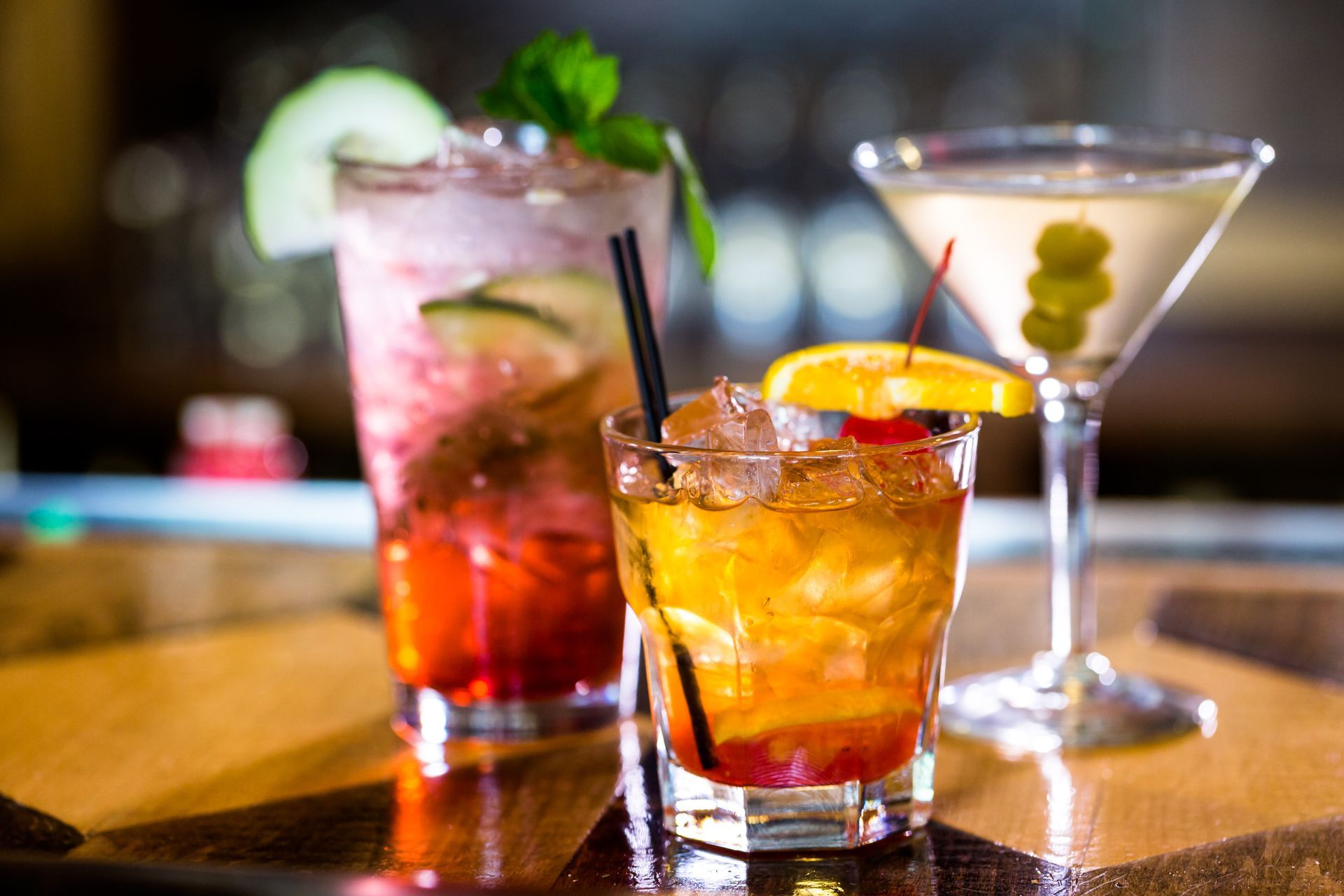 Three colorful cocktails on a bar, including a pink drink, amber drink, and martini with olives.