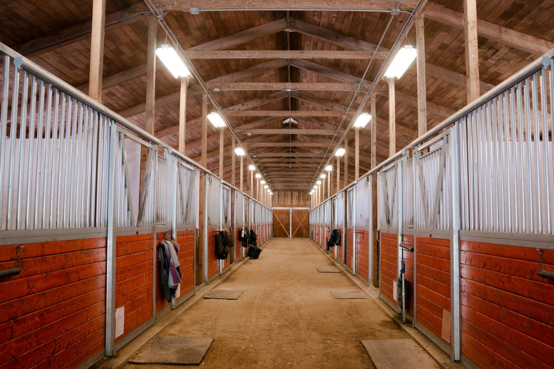 Rustic barn interior featuring weathered wood beams, illuminated by cage-style wall lights, with a tractor and barrel.