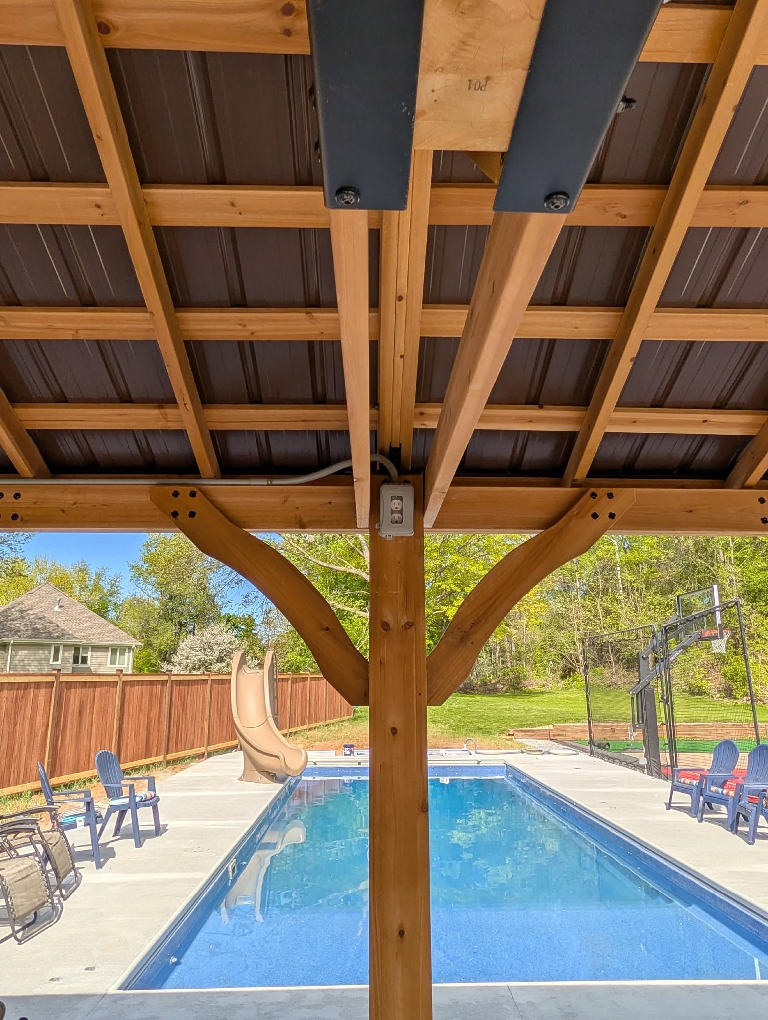 Covered swimming pool with blue water, lounge chairs, and wooden beams overhead in a backyard setting