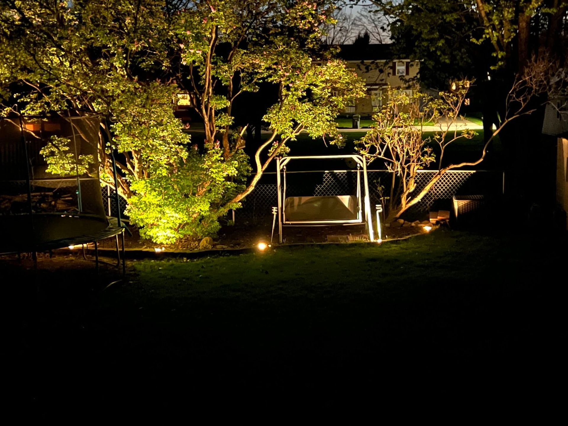 Illuminated garden patio at night with trees and a white swing bench