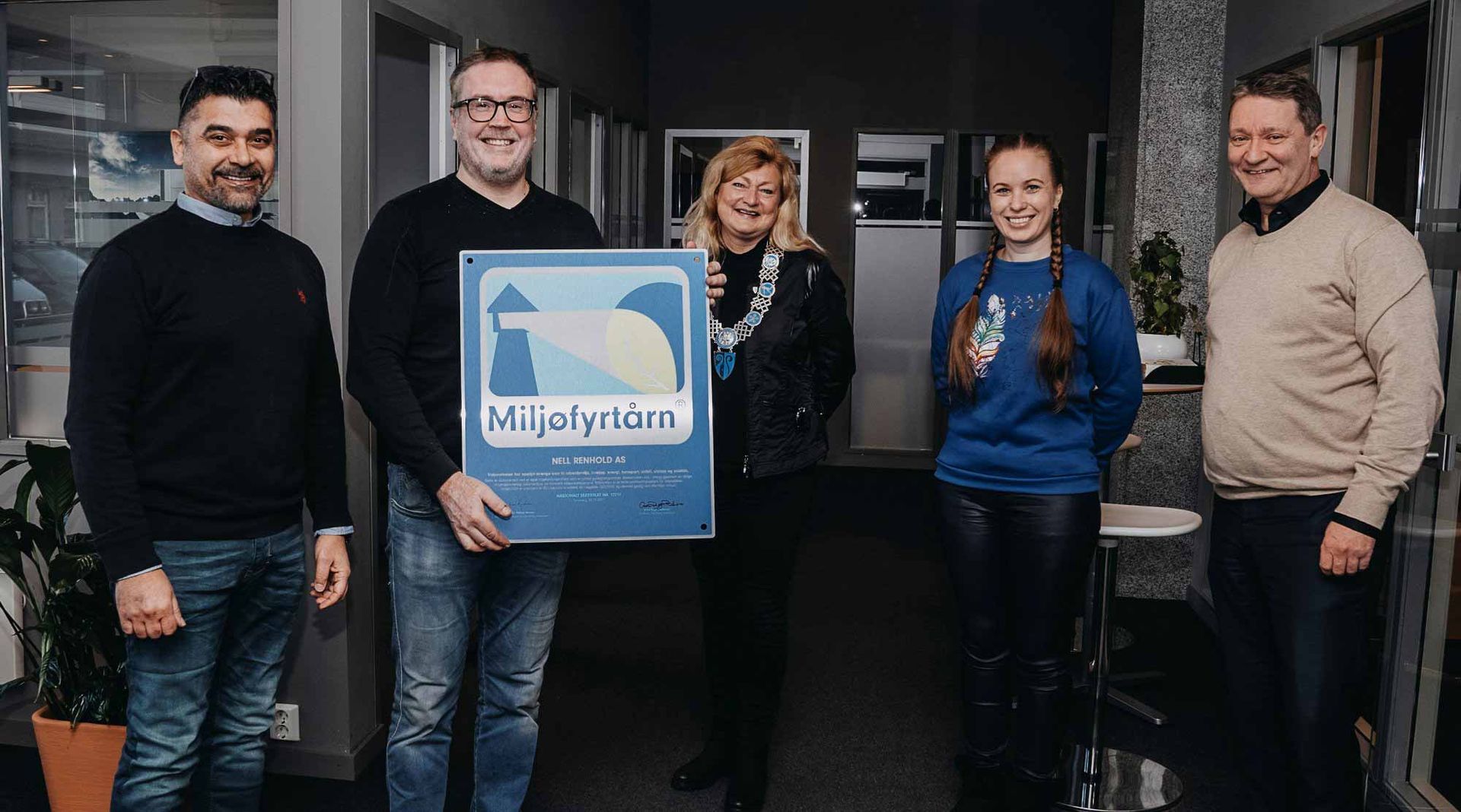 Man in glasses, patterned sweater, jeans seated in a gray armchair, next to a potted plant in a bright office space.