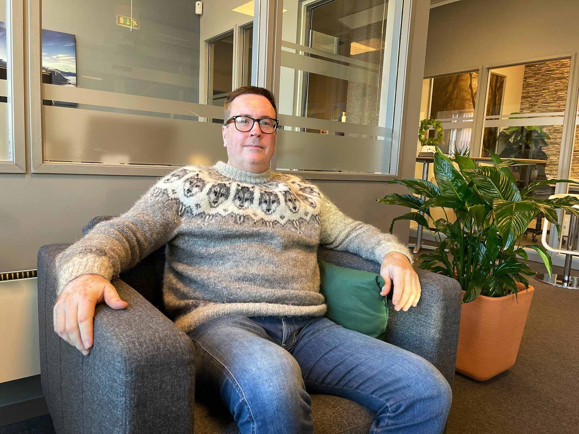 Man in glasses, patterned sweater, jeans seated in a gray armchair, next to a potted plant in a bright office space.