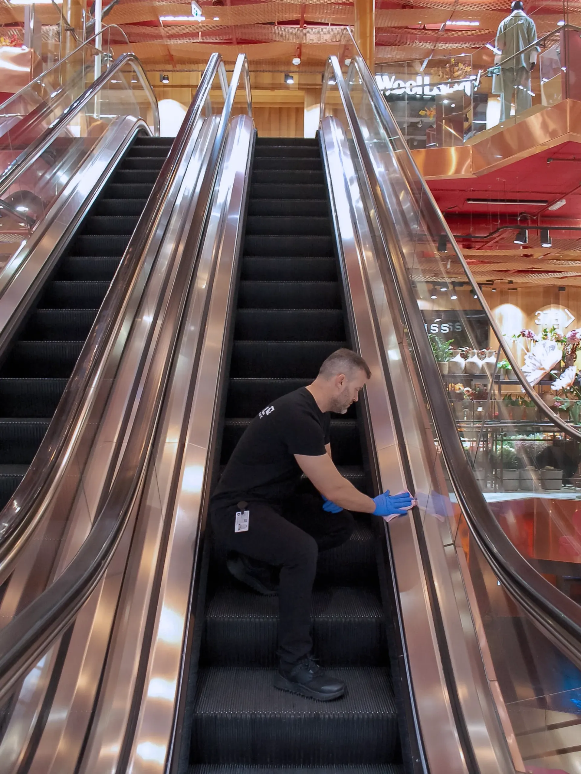 En person som rengjør en rulletrapp. De er på trappen og bruker et rengjøringsverktøy på sideskinnen. Lyse, moderne omgivelser.