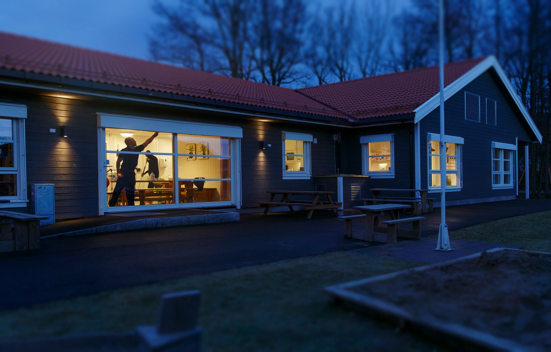 Mørkeblå bygning med rødt tak; innvendige lys på. Person synlig inne gjennom stort vindu. Skumring.