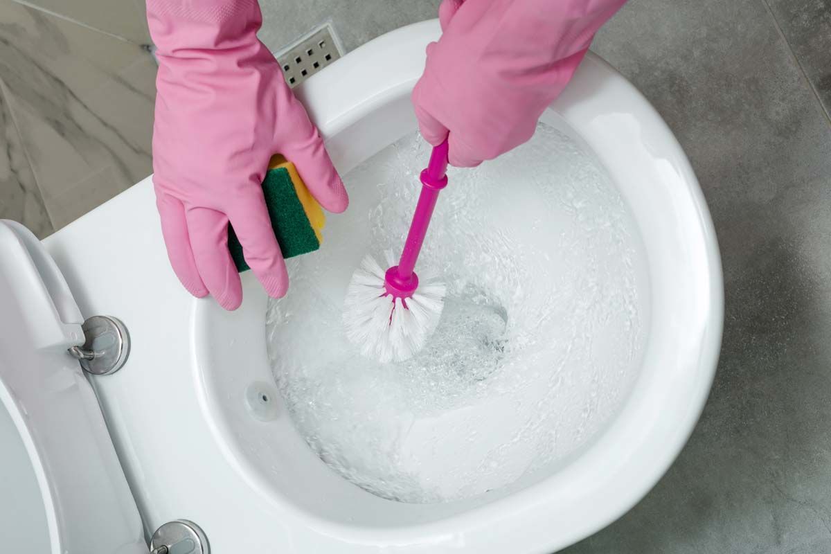 Person wearing pink gloves cleans a toilet with a brush and sponge, gray tiled floor.