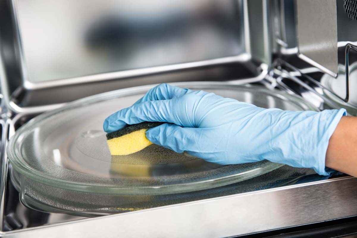 En person med blå hanske rengjør glassplaten i en mikrobølgeovn med en svamp.