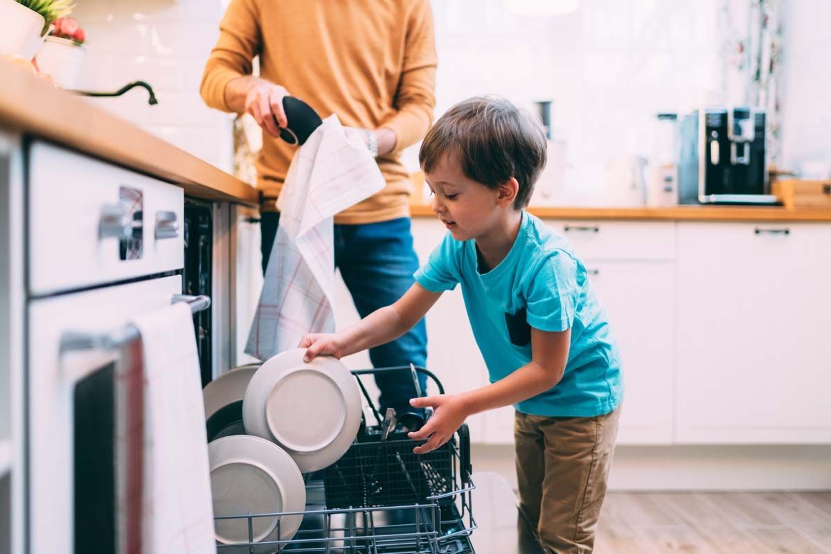Et barn og en voksen tømmer en oppvaskmaskin på et lyst kjøkken. Barnet plasserer tallerkener på en hylle.