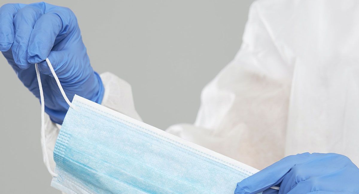 Person i blå hansker og hvit beskyttelsesdrakt som holder en blå ansiktsmaske i øreløkkene.