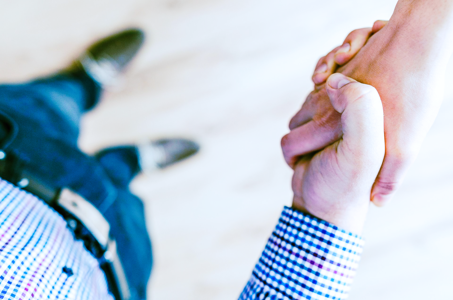 To personer som håndhilser, sett ovenfra. En mann i blå rutete skjorte og jeans. Brune sko i bakgrunnen.