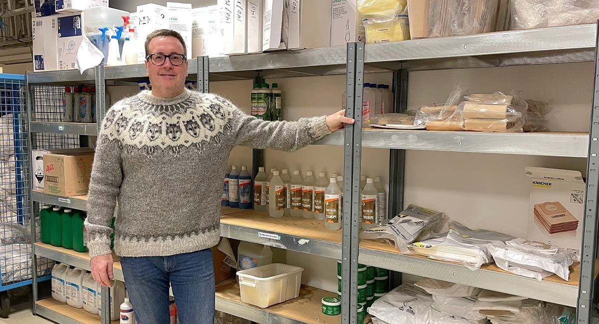 Man in sweater stands by shelves of supplies in a warehouse.
