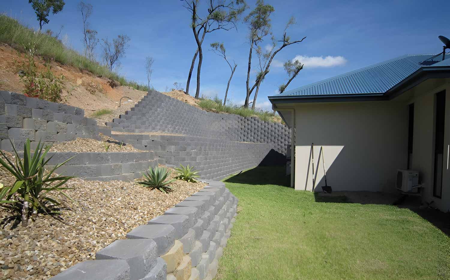 A House Is Sitting On Top Of A Grassy Hill Next To A Stone Wall — Austen Landscaping & Earthmoving in Deeragun, QLD