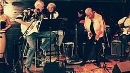 Musicians performing on a small stage with microphones, guitars, and drums under warm lighting.