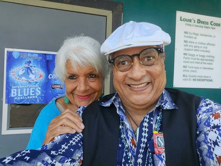 Two smiling people in blue patterned outfits, posing together outside a blues venue sign.