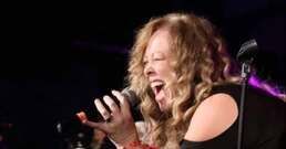 Singer with curly blonde hair passionately performing into a microphone on a dark stage