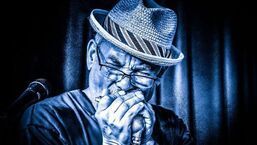 Blue-toned performer in a fedora playing harmonica on a microphone, lit by stage curtains
