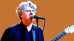 Musician singing into a microphone while holding a guitar, against an orange background.