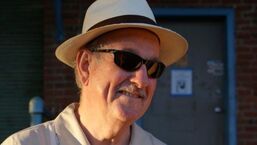 Man in a white hat and sunglasses smiling outdoors beside a brick building.