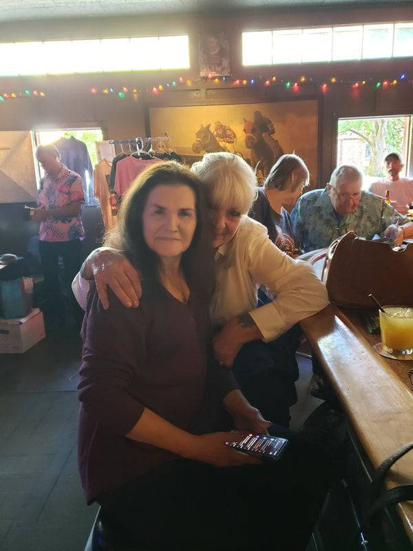 Two women sitting together at a table in a warmly lit room with string lights and other people in the background