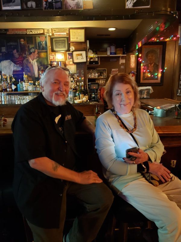 Two people seated in a warmly lit bar, smiling at the camera, with shelves and neon decor behind them.