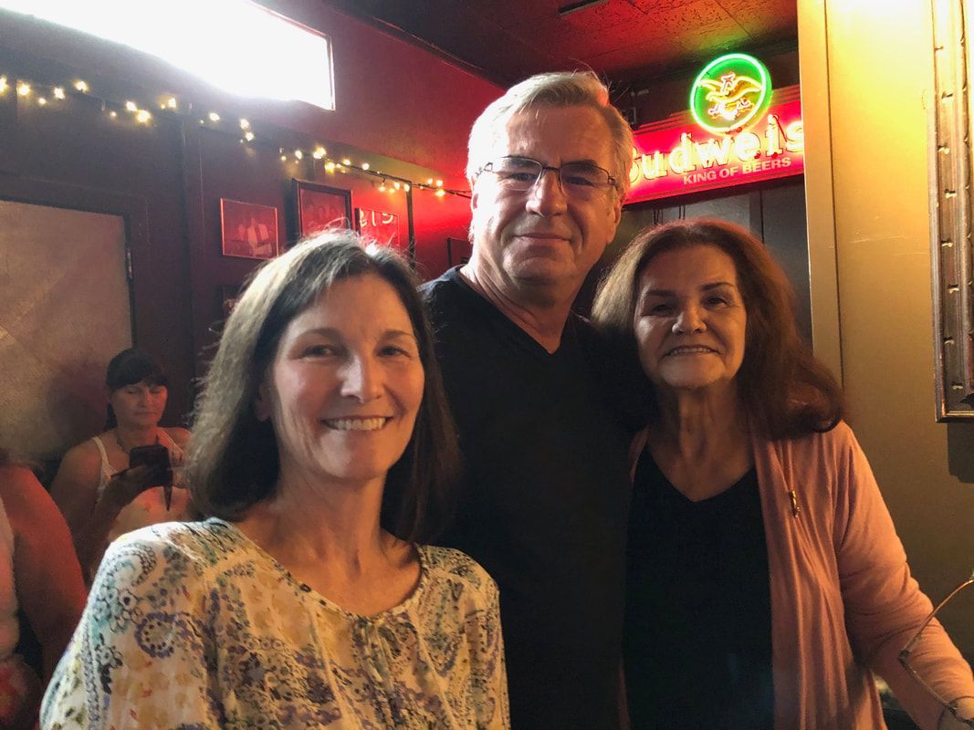 Three people smiling in a dimly lit bar with neon signs in the background.