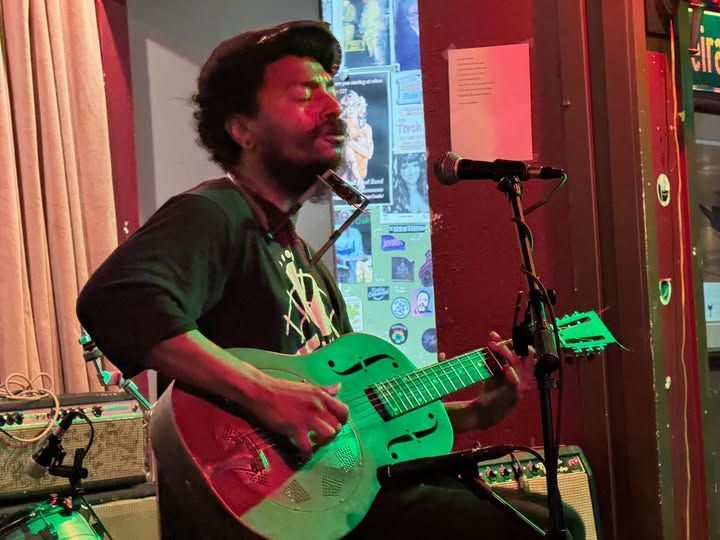 Musician playing green electric guitar and singing into a microphone on a small indoor stage