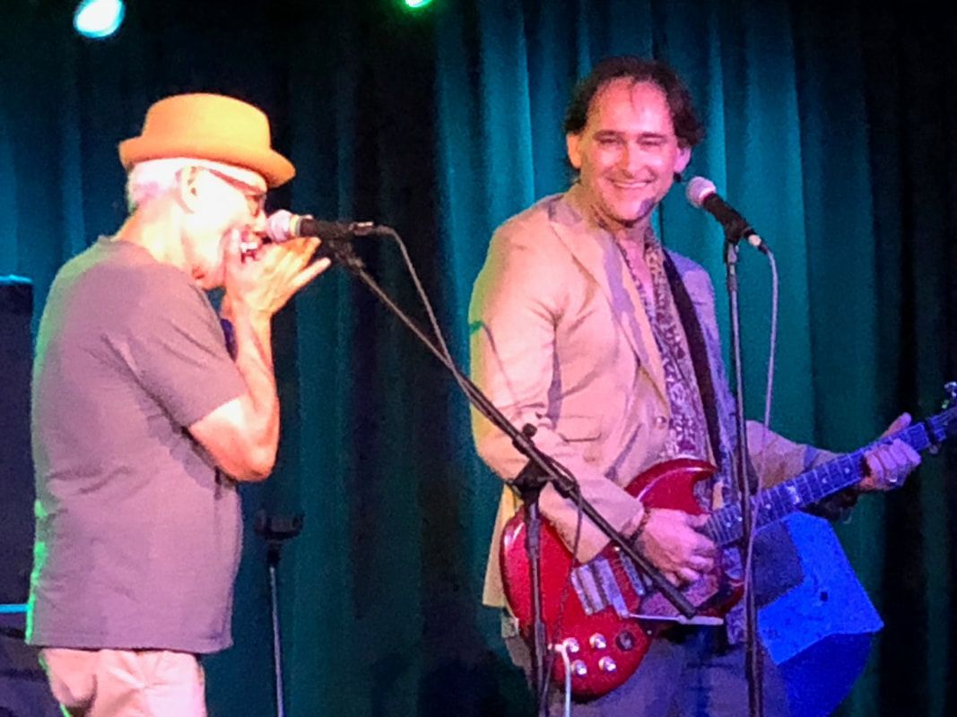 Two musicians performing on stage, one singing into a microphone and one playing a red electric guitar.