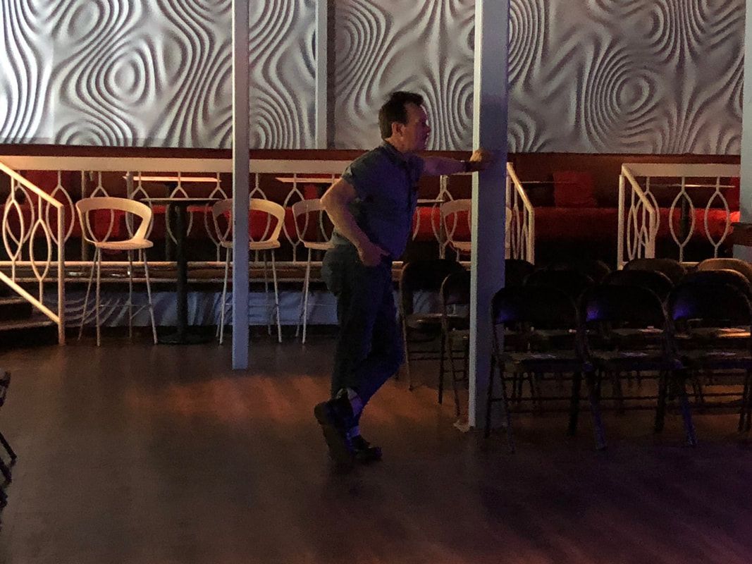 Man dancing in a dimly lit room with patterned walls and chairs along the back wall