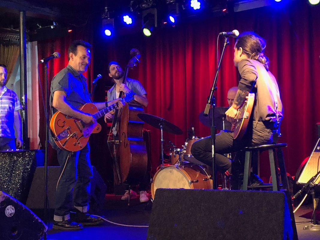 Live band performing onstage under blue and red lights, with guitars, drums, and red curtains.