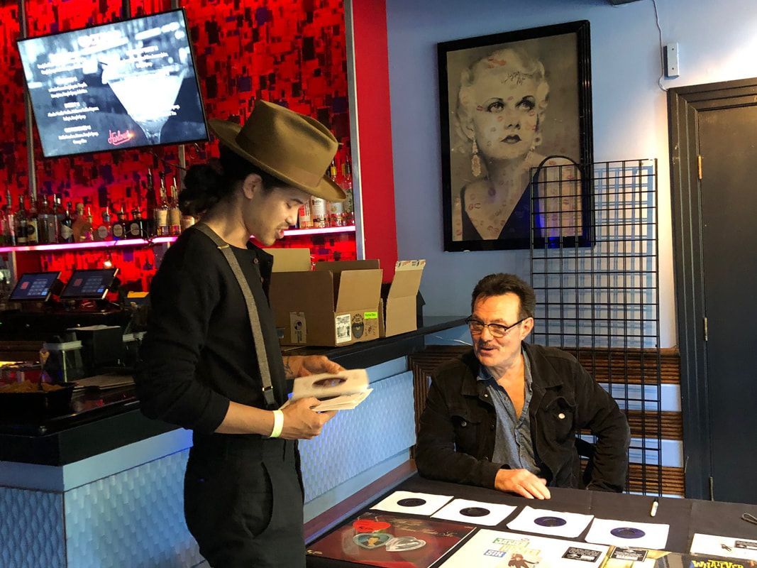 Two people in a red-lit gallery, one seated at a table and one standing with papers.