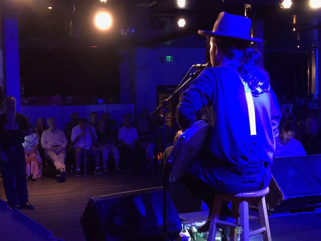Musician in a hat sits on a stool onstage under blue-purple lighting, facing an audience