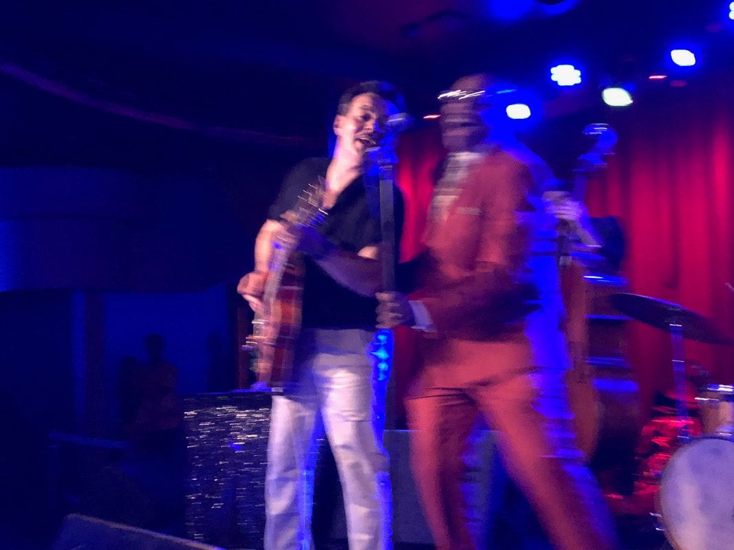 Two musicians performing onstage under blue and purple lights, one holding a guitar.