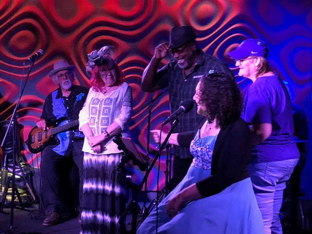 Band performing onstage under purple and red lights, with one guitarist and seated audience members nearby