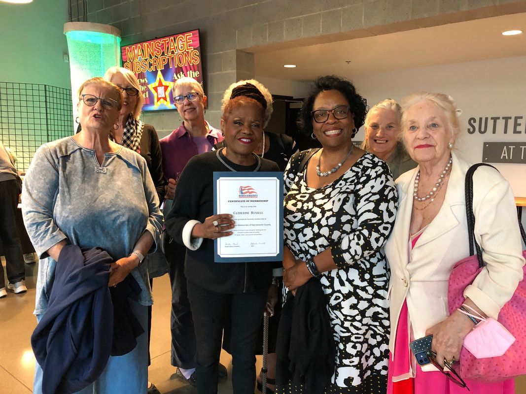 Five people posing indoors, one holding a framed certificate, with artwork and a sign in the background
