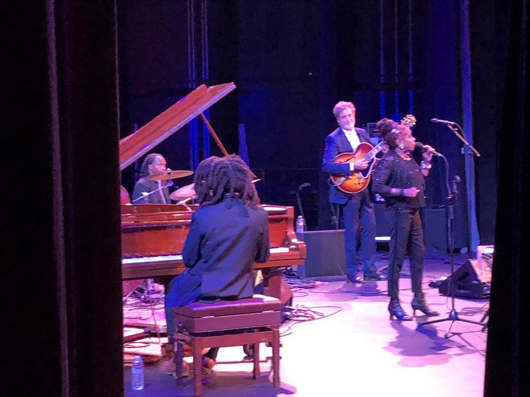Small band performing on a stage, with pianist at a grand piano and two vocalists at microphones.