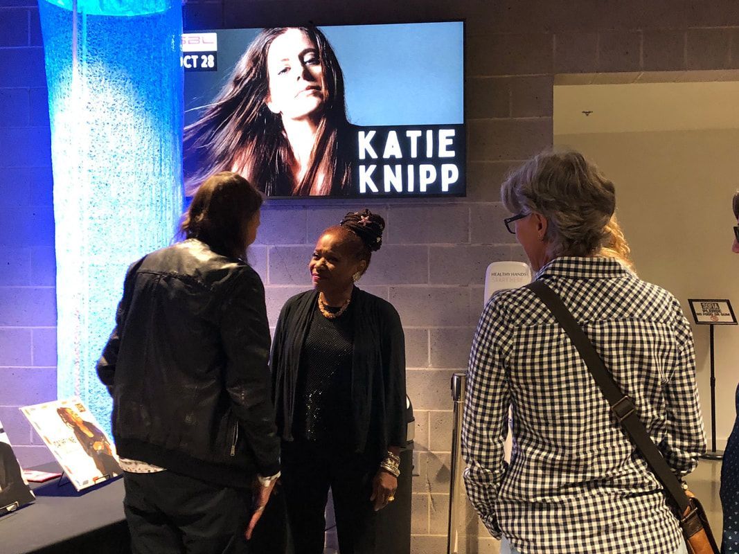 Three people standing in a lobby beneath a large “KATIE KNIPP” screen