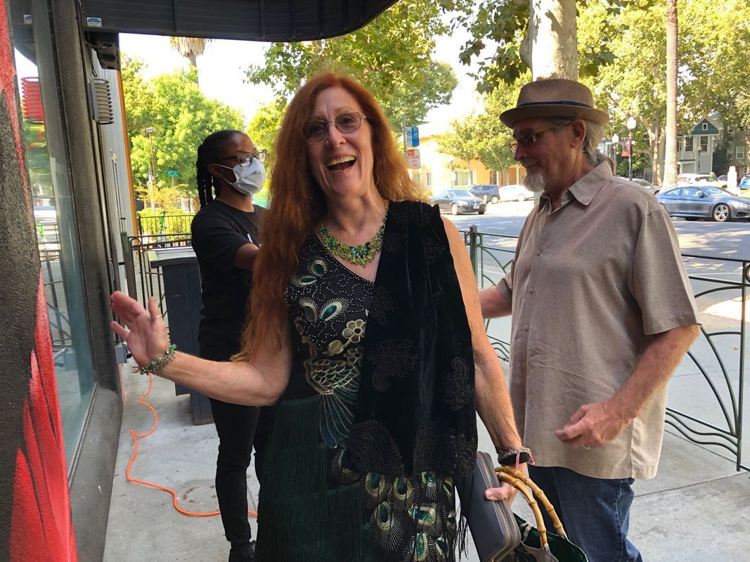 Smiling woman with long hair stands on a sidewalk, gesturing beside a storefront; man in hat walks behind her.