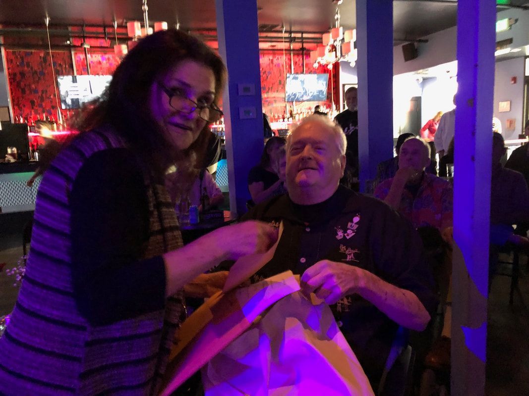 Two people smiling in a dimly lit bar, holding a large yellow bag under purple and red lights.