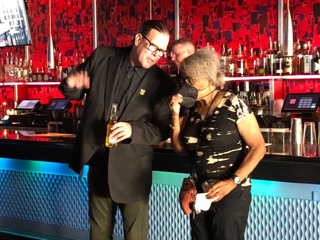 Two people posing at a red-lit bar, one holding a drink and the other leaning in with a smile.