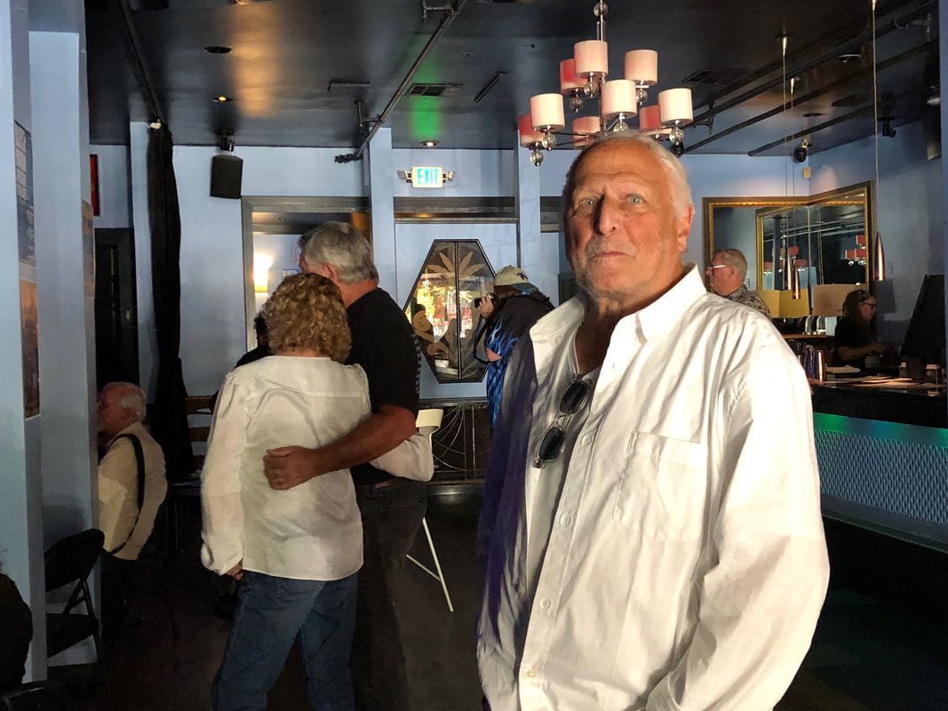 Man in a white shirt standing in a dimly lit indoor lounge with people in the background
