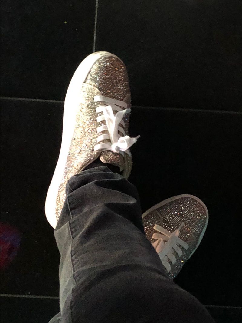 Two glittery sneakers with white laces, one foot crossed over the other on a dark floor.