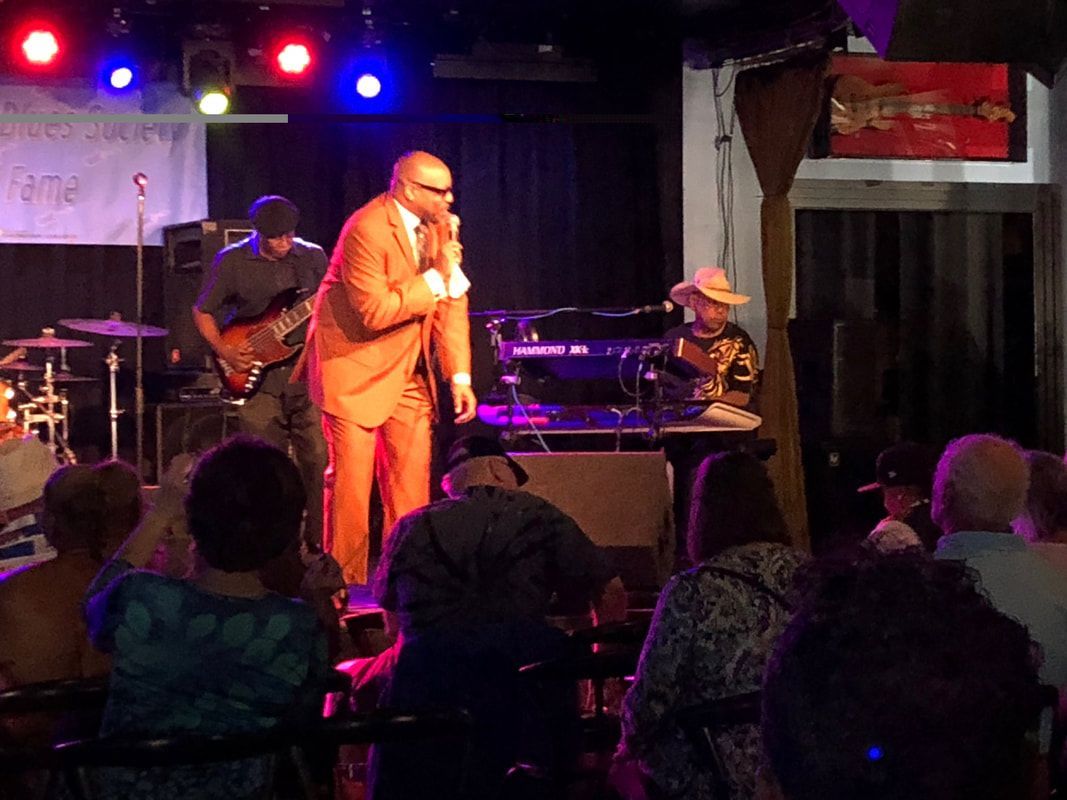 Singer in tan suit performing onstage under red and blue lights, with band and audience visible